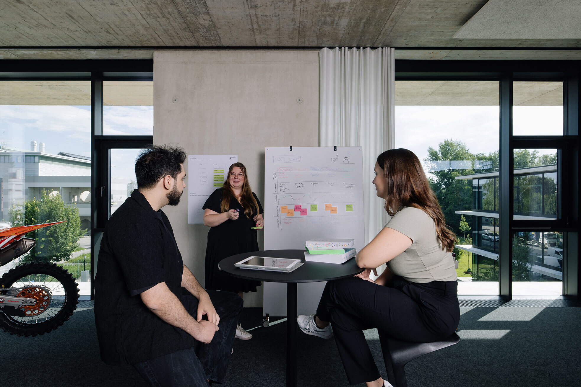 Three people in a modern office setting, collaborating at a round table with a flip chart displaying colorful notes. Large windows in the background.