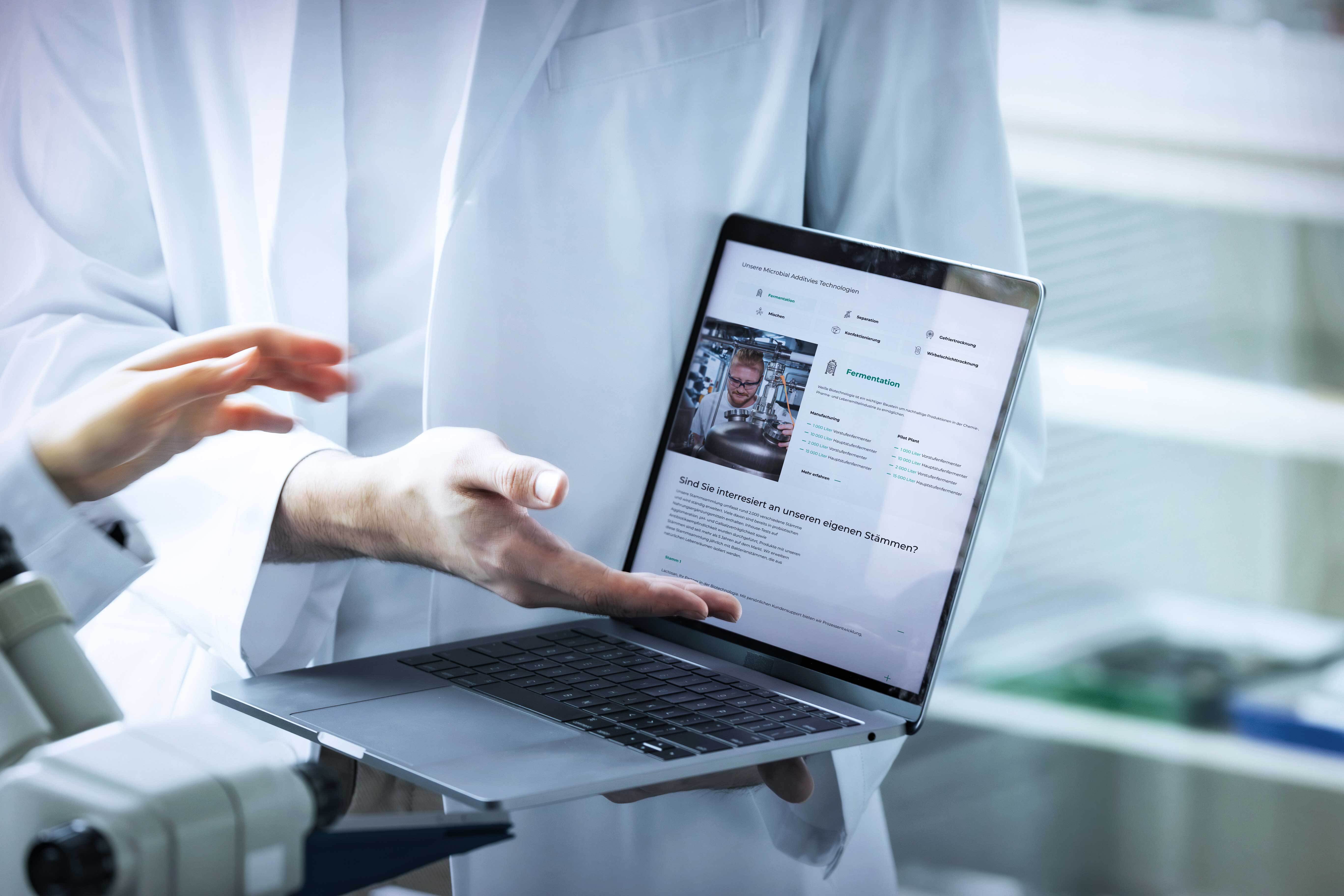Person in a lab coat holding a laptop displaying medical data, near a microscope in a laboratory setting.